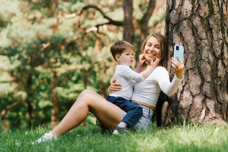 Happy mother and son in the park on a summer day taking a selfie, sitting near a tree on the grass