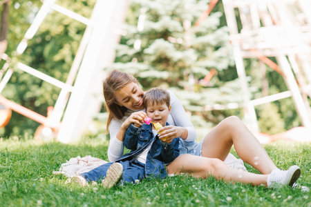 Happy and cheerful mom and son are blowing soap bubbles in a summer park, sitting on the grassの写真素材
