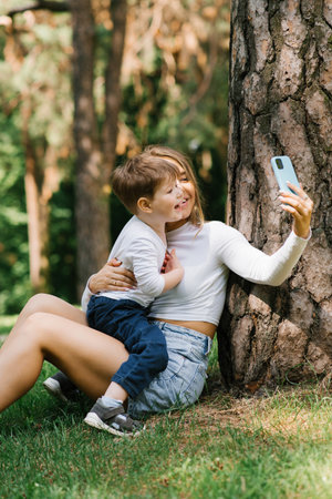 European family having fun, a mother and her son taking a selfie with a mobile phone in the park together, the concept of people of love and happinessの写真素材