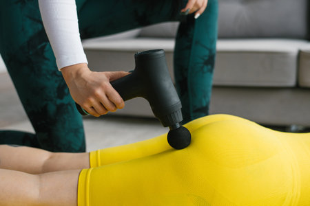 Female massage therapist does massage exercises on the hips with the help of a shock vibrator massager to a female client at home. Shock therapy for a restorative massage of the athletic bodyの写真素材