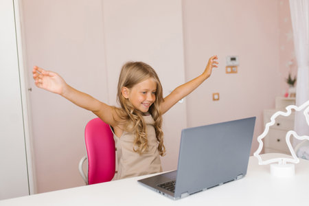Happy seven year old girl raises her hands to the sides while talking on a video chat on a laptop, sitting at a tableの写真素材
