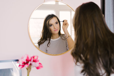 Girl with dark hair stands in front of a mirror and does makeup, holding a brush in her hand, creating an image, beauty and styleの写真素材