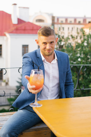 Young handsome man is sitting in an outdoor summer cafe in a blue suit and holding a cocktail glass in his handの写真素材