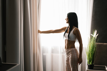 Brunette woman stands at the bedroom window in the morning. She has parted the curtains on the window and is looking at the streetの写真素材