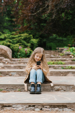 Caucasian girl of seven years old sits on stone steps in a park and communicates online with friends on a smartphoneの写真素材