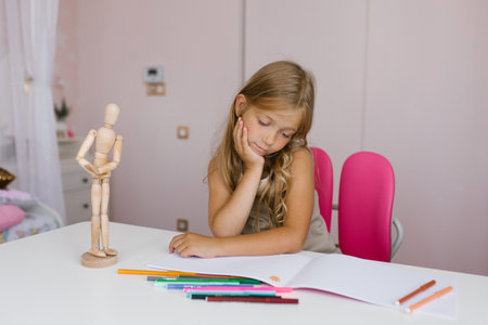 Seven year old schoolgirl is doing homework at her desk at home or at school, drawing with felt-tip pens, looking thoughtfully into a notebookの写真素材