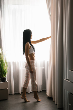 Single woman is a beautiful Caucasian woman in pajamas without standing at the window of a house or in a hotel room and looking at the street on a sunny dayの写真素材