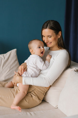Happy beautiful young mother holds her little son in her arms on the sofa and looks at him with love...