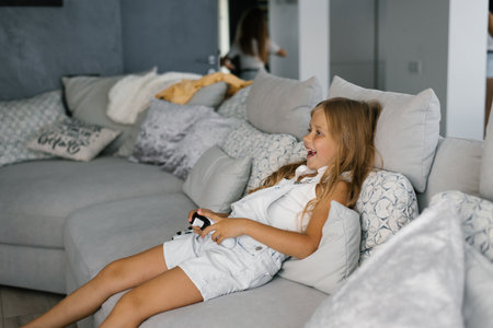 Child girl plays a video game console using a joystick or controller, sitting at home on the couch, the concept of leisure for real peopleの写真素材