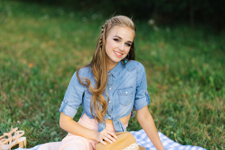 Pretty caucasian young woman with long hair is sitting on a blanket in nature on a sunny summer day....