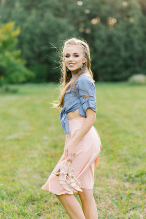 Young Caucasian woman or girl in a summer dress and a denim shirt is walking on the grass, holding shoes in her hands, relaxing outside the city on a weekend in summerの写真素材