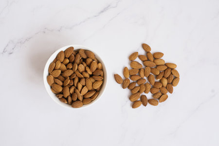 Almonds lie in a white bowl and are scattered on a marble table.の写真素材