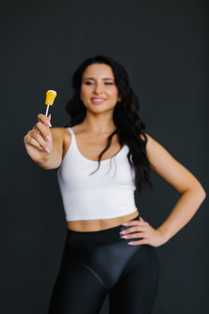 Young female sports fitness trainer holding lollypop in outstretched hand on black background