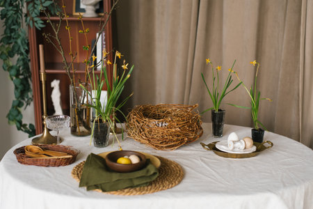 Table with a white tablecloth and a basket of easter eggs. There are also some flowers and a bird in...