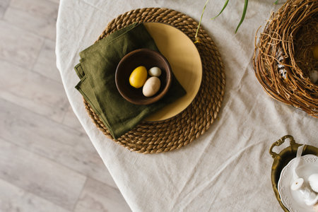 Table with a green napkin and a yellow plate with easter eggs and a bowl. The table is set for a mealの写真素材