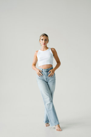 Young woman is wearing a white tank top and blue jeans. She is standing in front of a white backgroundの写真素材