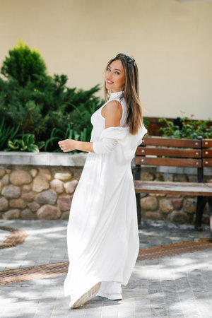 Happy woman in a white dress is standing in front of a bench. She is smiling and looking at the camera. The bench is located near a buildingの写真素材
