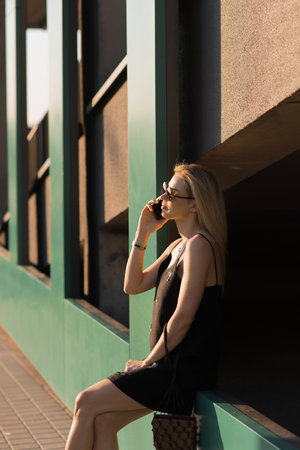 Young woman in a black dress, talking on a cell phone, walking on a city street in the summertimeの写真素材