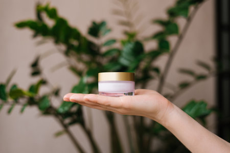 Small jar of cream on a woman's hand against a background of a green plant. The jar is transparent, with a gold lid. The concept of organic cosmeticsの写真素材