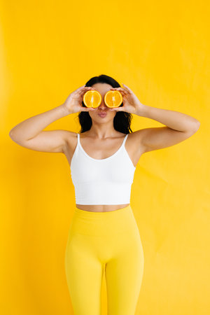 Young woman covers her eyes with half oranges on yellow background. The concept of healthy food. Skin care and beautyの写真素材