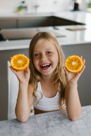 Child girl with oranges in her hands sits at a table and laughs. Healthy vitamin food. Funny girl with fruitの写真素材