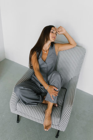 Sad woman is sitting in a chair wearing a gray vest and gray pants. She is wearing a necklace and has her hand on her headの写真素材