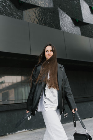 Caucasian woman wearing a black jacket and white pants walks down a street with carrying a black purse and long hairの写真素材