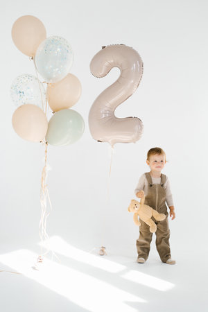 Boy child stands in front of birthday balloons with the number two. The child is holding a stuffed animal teddy bearの写真素材