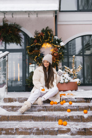 Young woman is sitting on the stairs with tangerines scattered around her in her hand. Cozy and warm atmosphere before Christmasの写真素材