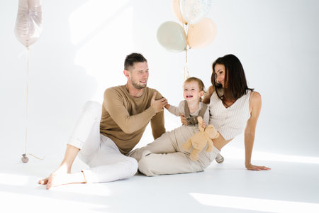 Mom, dad and a two-year-old birthday boy are sitting on the floor near a bunch of festive balloons and having fun togetherの写真素材
