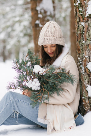 Young woman is sitting in snow with white hat on and holding bouquet of pine branches and cottonsの写真素材