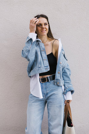 Young woman in a blue jacket and jeans smiles and holds a purse in her hands against a beige wall in the cityの写真素材