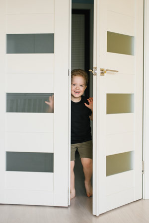 Child boy is standing in a doorway, looking out into the hallway. He is wearing a black shirt and shortsの写真素材
