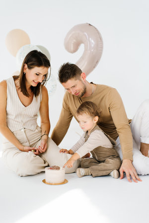 Young family with a child is sitting on the floor in front of a birthday cake in honor of the second birthday with balloons and the number twoの写真素材