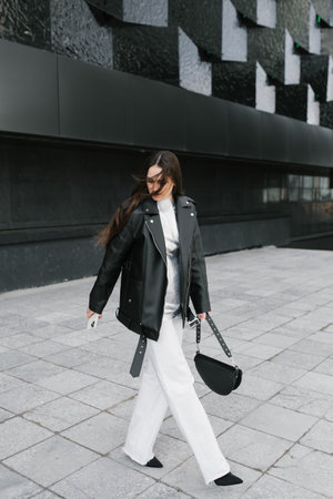 Stylish woman wearing a black leather jacket and white pants walks down a sidewalkの写真素材