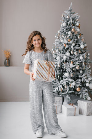 Children girl is standing in front of a Christmas tree, holding a gift box. Concept of joy and excitement, as likely preparing to give the gift to someone specialの写真素材