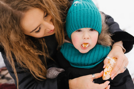 Young mom is holding a child in her arms and feeding him a piece of orange at winter outdoor
