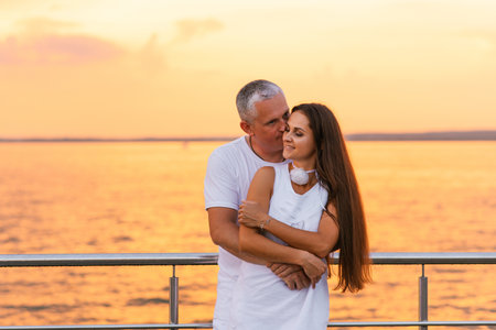 Happy couple is hugging on pier by the water. Young woman has long hair and the man is kissing her. Scene is romantic and peacefulの写真素材