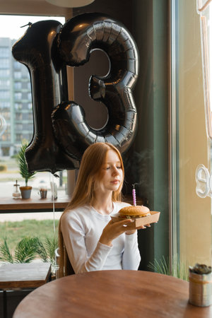 Teenager girl is sitting at a table with a birthday cake and a balloon number thirteen. She is holding a candle and is about to blow it outの写真素材