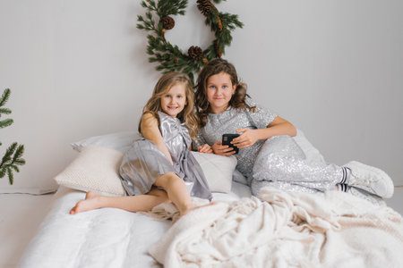 Two young sisters girls are sitting on bed, one of them holding smartphone. The room is decorated with Christmas trees and pine cones, giving it a festive atmosphereの写真素材