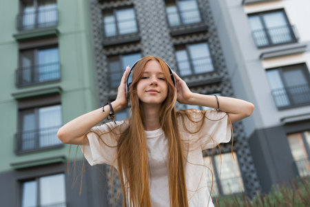 Teenager with long red hair is wearing headphones and smiling, standing in front of a urban buildingの写真素材