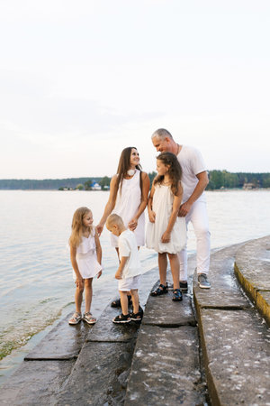 Happy family of five is standing on stone staircase by a body of water. The children are wearing white dressesの写真素材