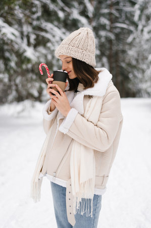 Young caucasian woman in white coat, hat is holding cup of coffee and candy cane. She is enjoying the winter weather and the warmth of her drinkの写真素材