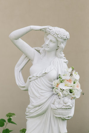 A graceful white statue adorned with a floral crown, holding a delicate bouquet of pastel flowers against a neutral background.の写真素材