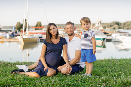A happy family with a pregnant mother, father, and child sitting on grass near a marina, surrounded by boats and water, under a clear blue sky.の写真素材