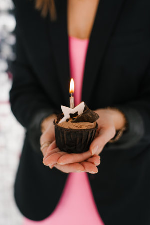 Close up of hands holding a chocolate cupcake with a burning candle on top, symbolizing celebration...