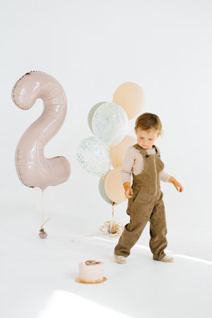 Birthday child boy stands in front of a festive cake and balloons, with a large balloon in the background that says twoの写真素材