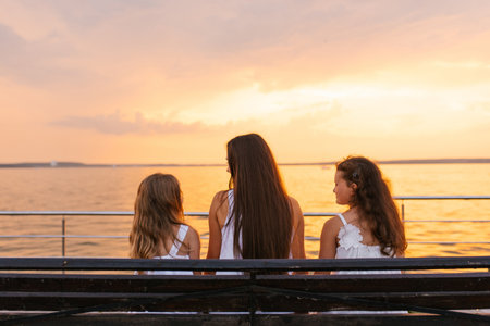 Mother sits on bench with two young girls near body of water, and the sky is a beautiful orange...