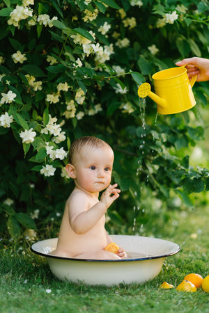 A baby sitting in a small tub outdoors, playing with an orange and water, while a yellow watering...