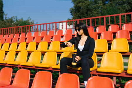 A woman in black sportswear holding a yellow shaker bottle, sitting confidently on colorful stadium seats under sunlight.の写真素材